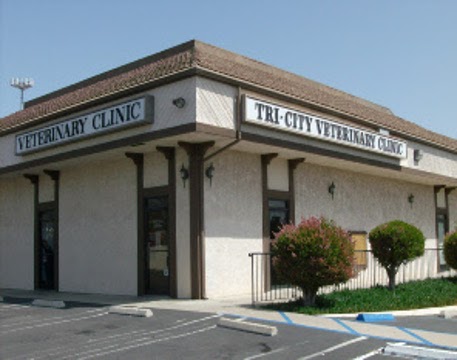 Corner building with parking spots visible, bushes on the right hand side as well as signage above both faces of the building. Side on the left is written in dark color text on a white backdrop and reads "Veterinary Clinic". Right side sign is also dark text on a white backdrop but reads "Tri-City Veterinary Clinic"
