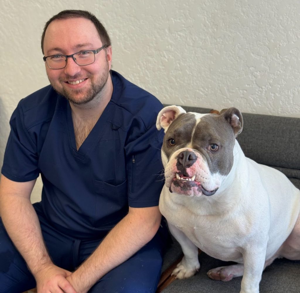 Man with short brown hair, a light beard and glasses in navy blue scrubs sitting next to a grey and white dog